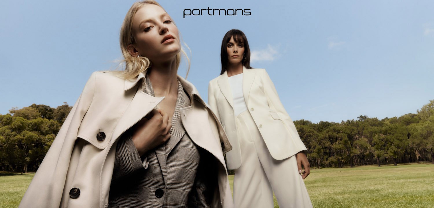 Two female models posing outdoors in a park with a grassy lawn and trees under a clear blue sky, wearing stylish coats.